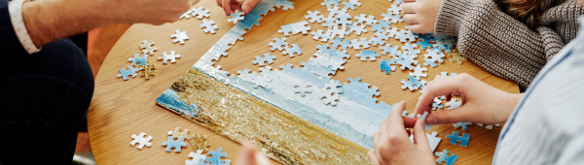 A group of people sat around a table doing a jigsaw puzzle of a beach scene