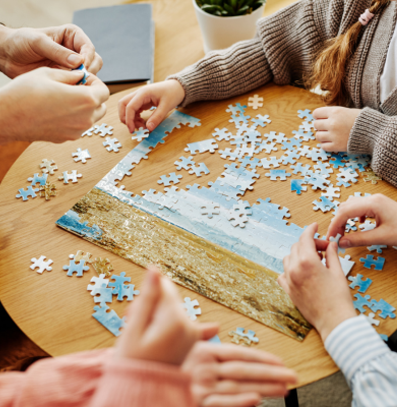 A group of people sat around a table doing a jigsaw puzzle of a beach scene