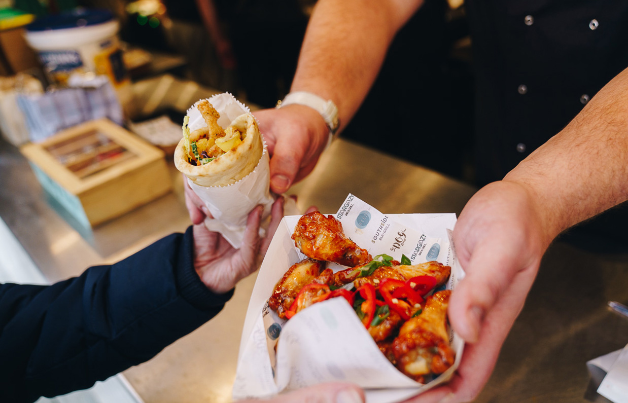 Two people's hands passing a gyros and selection of chicken legs
