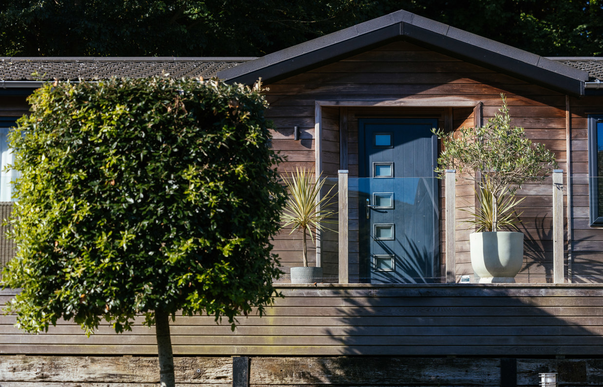 A holiday lodge with decking, plants and trees, surrounded by woodland