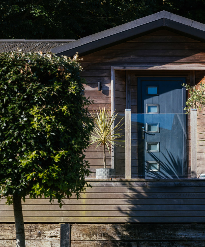 A holiday lodge with decking, plants and trees, surrounded by woodland