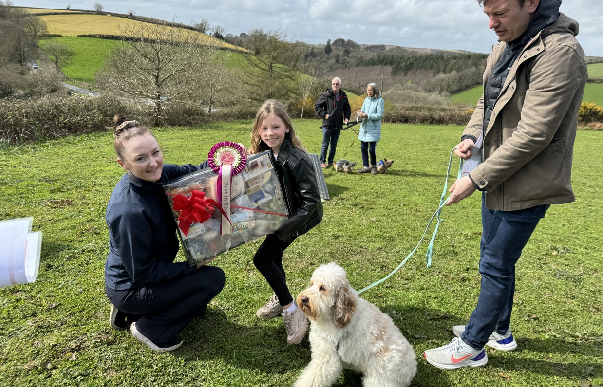 A small cream fluffy dog sat beside a man and a young girl being presented a prize by a woman for winning in Trufts Dog Show