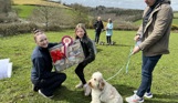 A small cream fluffy dog sat beside a man and a young girl being presented a prize by a woman for winning in Trufts Dog Show