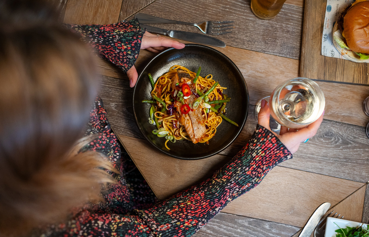 A salmon noodle bowl and a glass of wine by a woman on a table taken from above