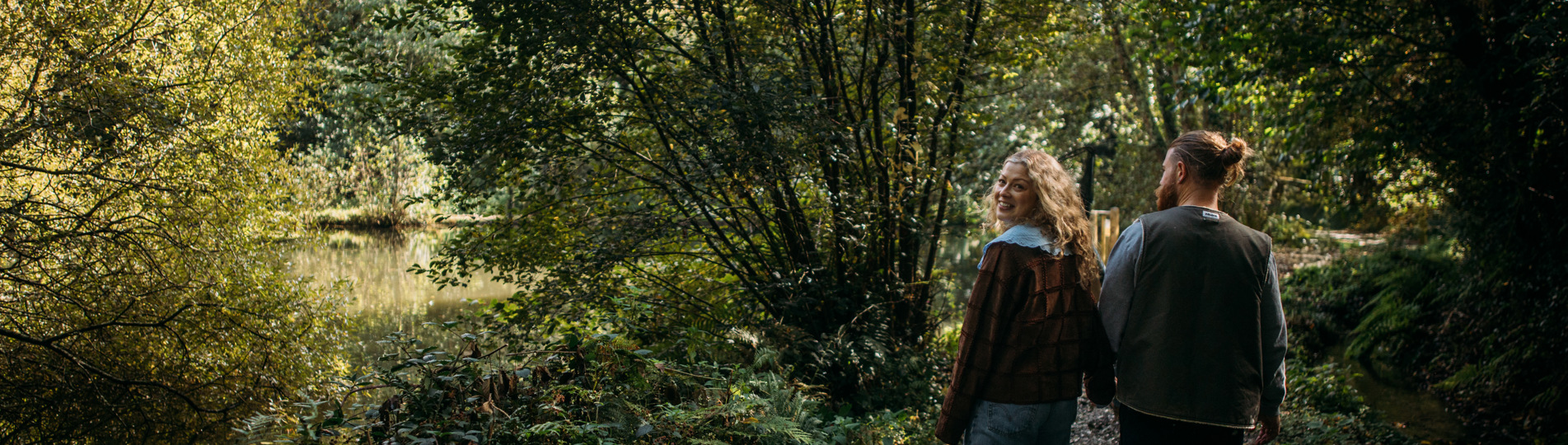 A couple walking through woodland together holding hands as the woman looks back towards the camera