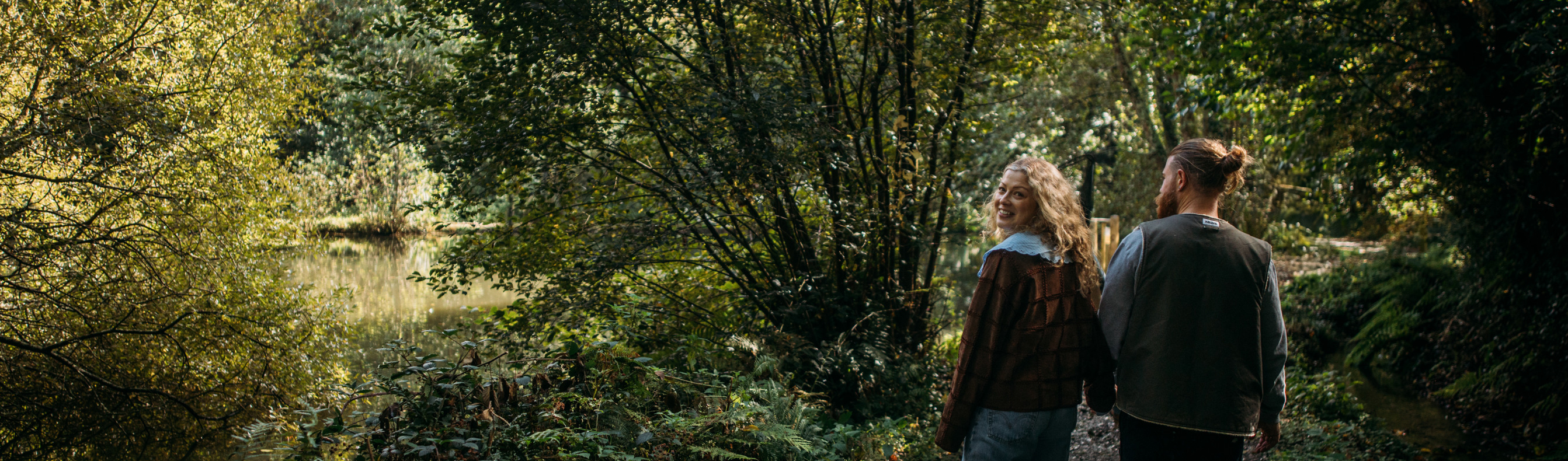 A couple walking through woodland together holding hands as the woman looks back towards the camera