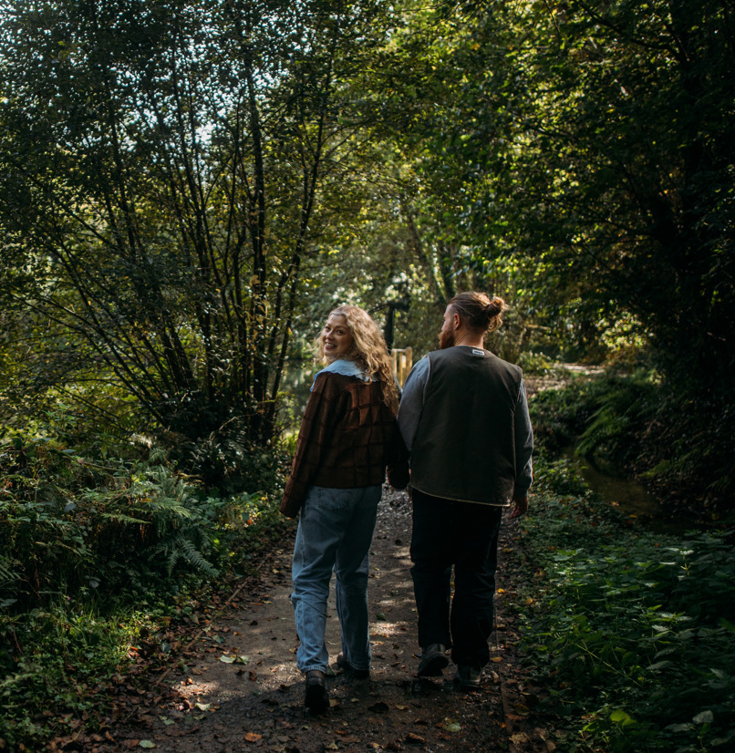 A couple walking through woodland together holding hands as the woman looks back towards the camera