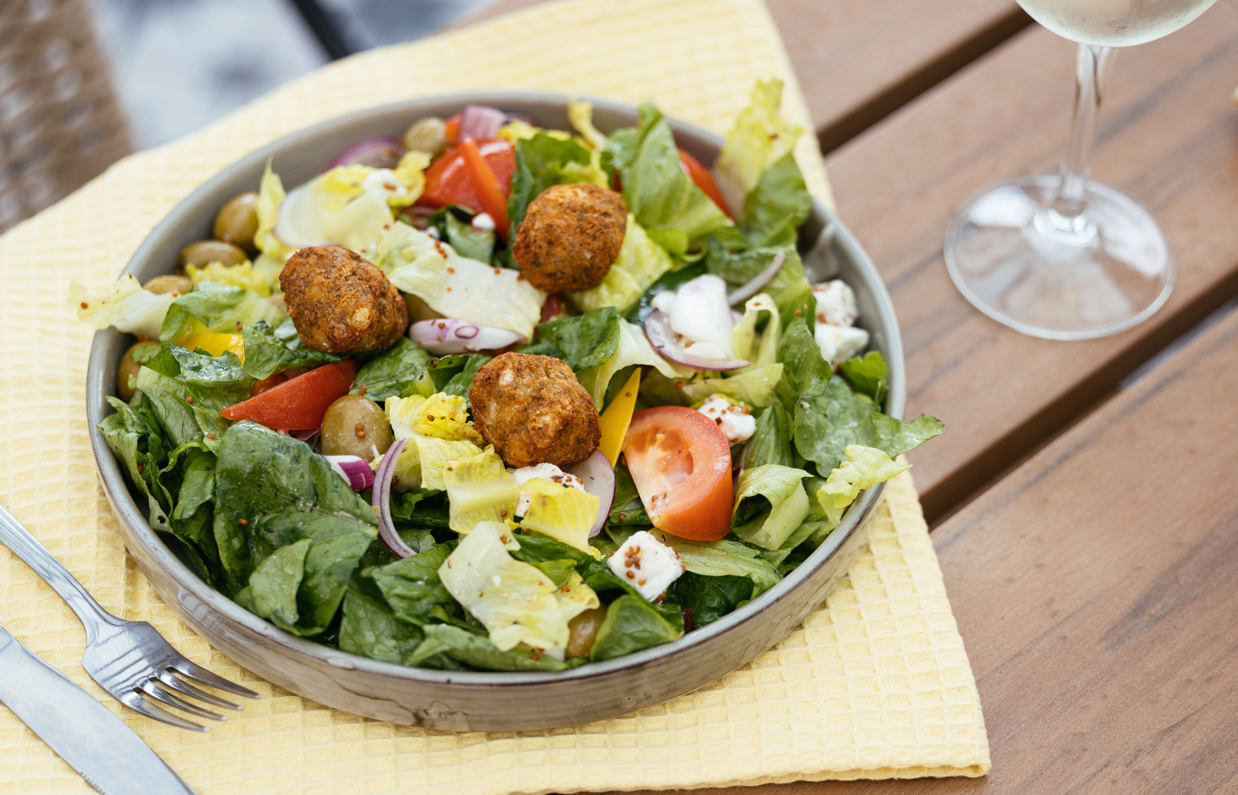 A leafy salad with falafel, tomato, olives and more on an outdoor table with a glass of white wine