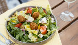 A leafy salad with falafel, tomato, olives and more on an outdoor table with a glass of white wine