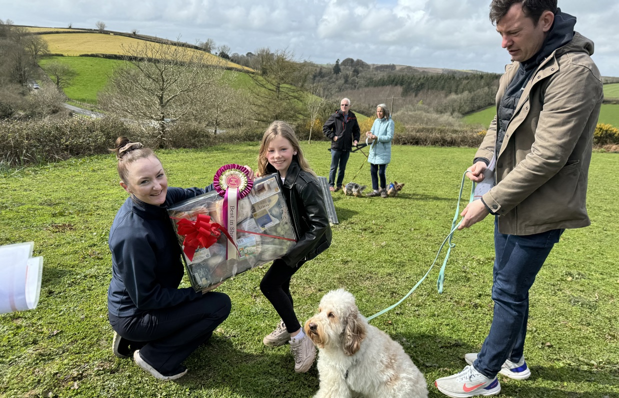 A small cream fluffy dog sat beside a man and a young girl being presented a prize by a woman for winning in Trufts Dog Show