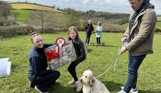 A small cream fluffy dog sat beside a man and a young girl being presented a prize by a woman for winning in Trufts Dog Show