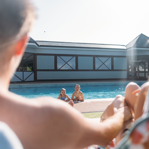 A young girl and boy in an outdoor pool with a mum and dad sat on sun loungers in the sun