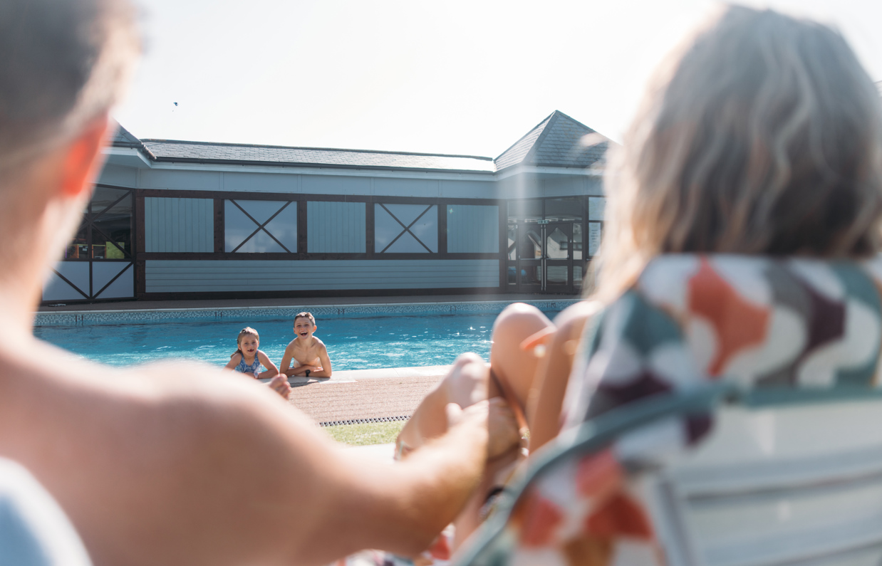 A young girl and boy in an outdoor pool with a mum and dad sat on sun loungers in the sun