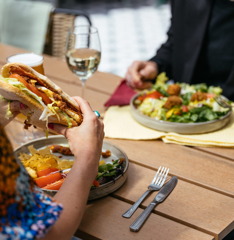 Two people sat outside eating hearty food dishes with a white wine and a pint of beer