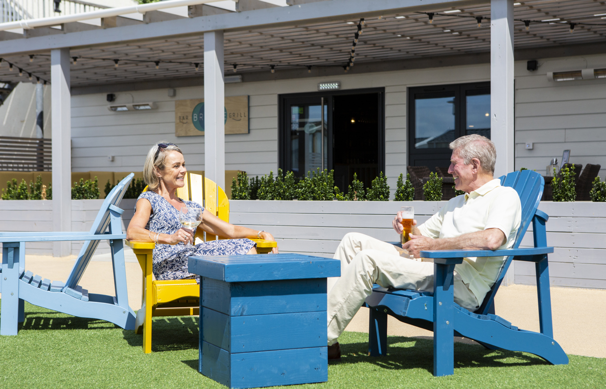 A couple sat outside Breeze Bar & Grill on a green area smiling at each other with drinks in hand