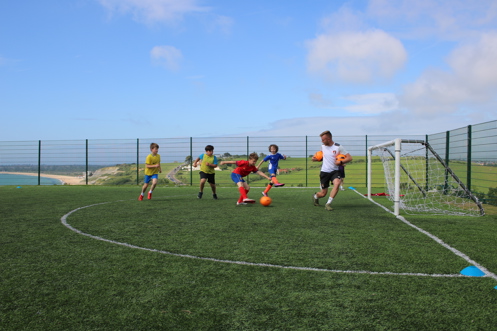 A group of young boys playing football at a sports field atop a hill overlooking the sea