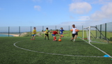 A group of young boys playing football at a sports field atop a hill overlooking the sea