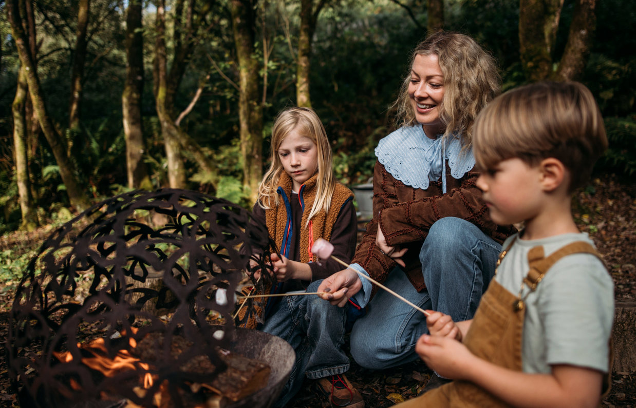 A mother and two young boys toasting marshmallows outside on a firepit
