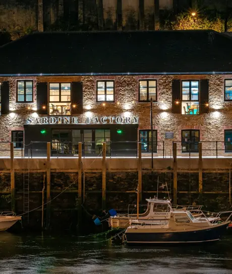 The external of The Sardine Factory restaurant lit up at night alongside the water with boats