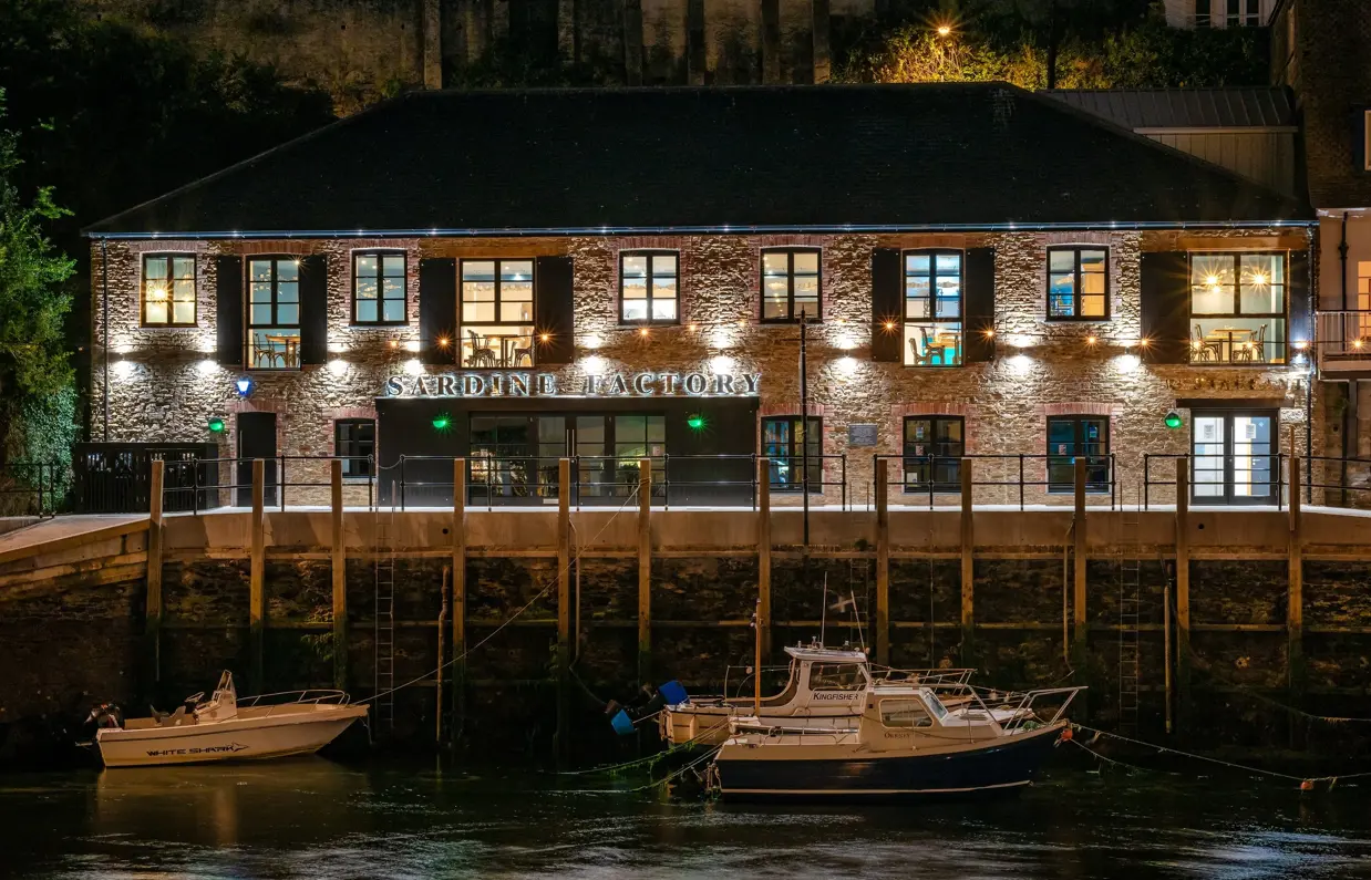 The external of The Sardine Factory restaurant lit up at night alongside the water with boats