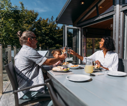 A couple with a young girl sat at an outside table of a luxury lodge on a sunny, blue sky day eating a meal outside