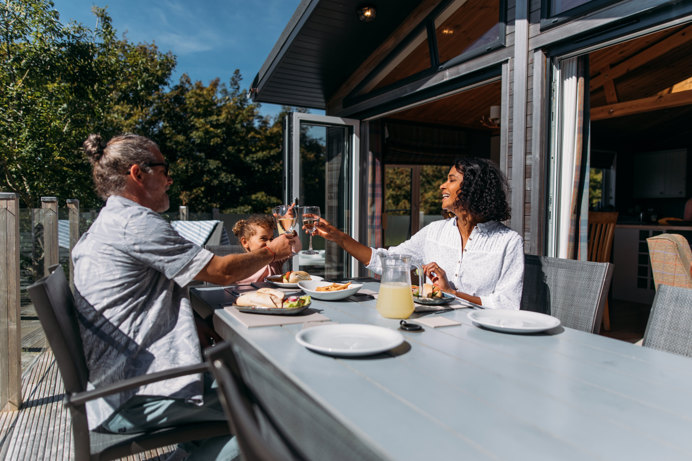 A couple with a young girl sat at an outside table of a luxury lodge on a sunny, blue sky day eating a meal outside