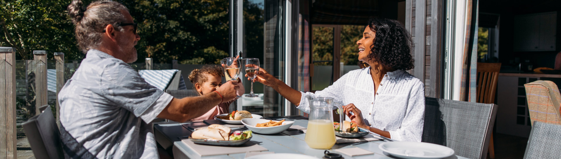 A couple with a young girl sat at an outside table of a luxury lodge on a sunny, blue sky day eating a meal outside