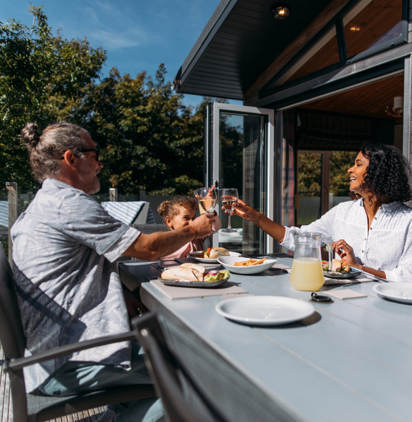 A couple with a young girl sat at an outside table of a luxury lodge on a sunny, blue sky day eating a meal outside