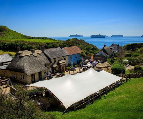 Aerial view of a quaint cottage-style pub with a thatched roof on the coast with countryside and sea views with cruise ships in the distance