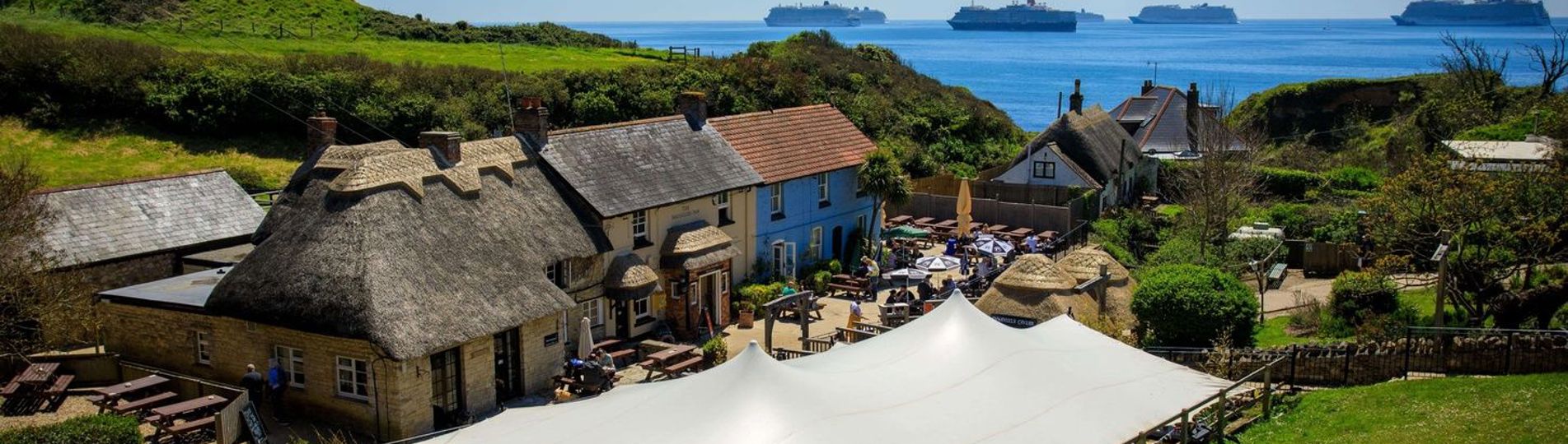 Aerial view of a quaint cottage-style pub with a thatched roof on the coast with countryside and sea views with cruise ships in the distance