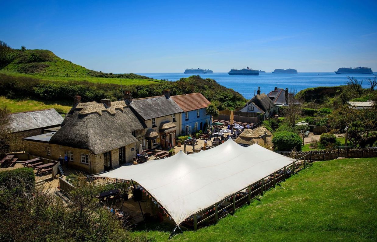 Aerial view of a quaint cottage-style pub with a thatched roof on the coast with countryside and sea views with cruise ships in the distance