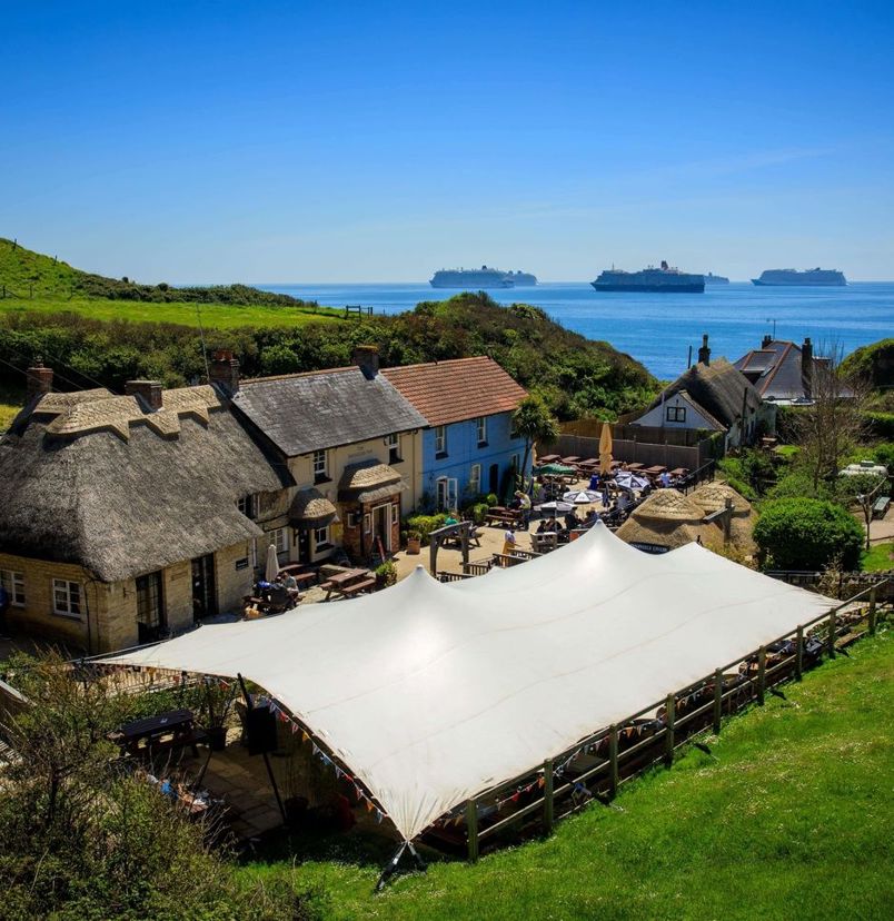 Aerial view of a quaint cottage-style pub with a thatched roof on the coast with countryside and sea views with cruise ships in the distance