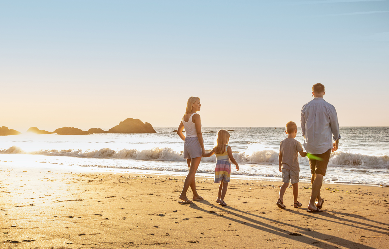 A family of four walking along the beach by the sea at sunrise with rocky mounds in the distance