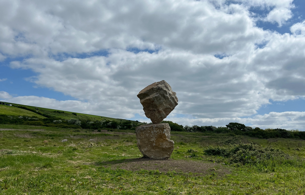 A carefully balanced stone sculpture in a countryside field with blue skies and clouds in the background