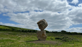 A carefully balanced stone sculpture in a countryside field with blue skies and clouds in the background
