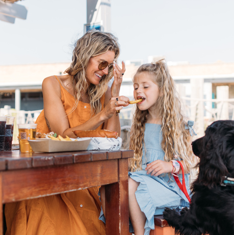 A young woman feeding a young girl a chip at a table outside whilst a black dog watches intently