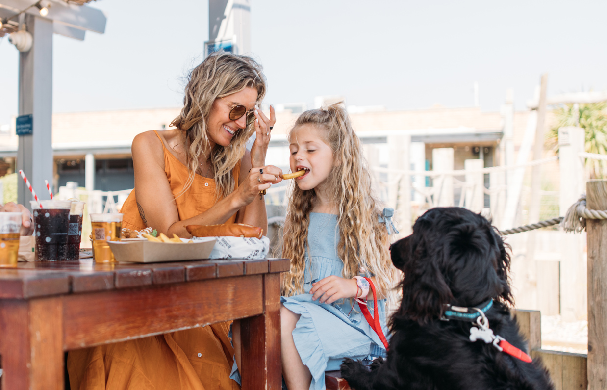 A young woman feeding a young girl a chip at a table outside whilst a black dog watches intently