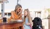 A young woman feeding a young girl a chip at a table outside whilst a black dog watches intently
