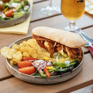 A chicken sandwich with crisps and salad on an outdoor table with a pint of beer and another salad dish