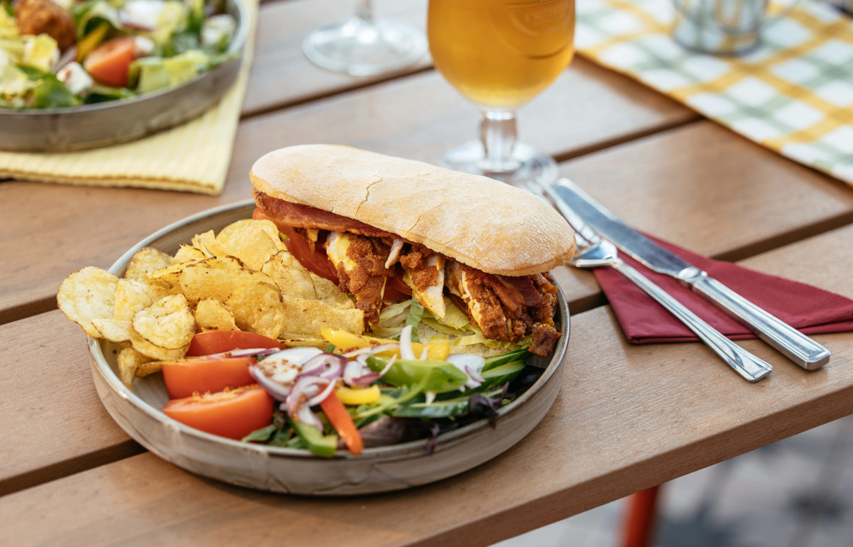 A chicken sandwich with crisps and salad on an outdoor table with a pint of beer and another salad dish