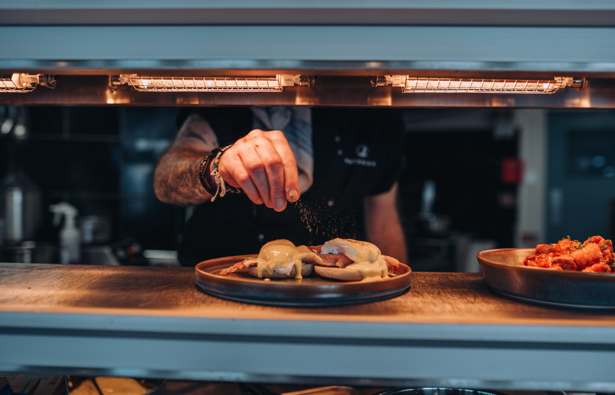 A plate of food in the serving counter of Drift Bar & Grill having the finishing touches and seasonings added to it by a chef in the kitchen