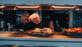 A plate of food in the serving counter of Drift Bar & Grill having the finishing touches and seasonings added to it by a chef in the kitchen