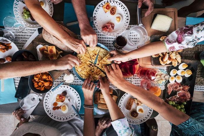 A selection of food dishes on a table with various people reaching for different items