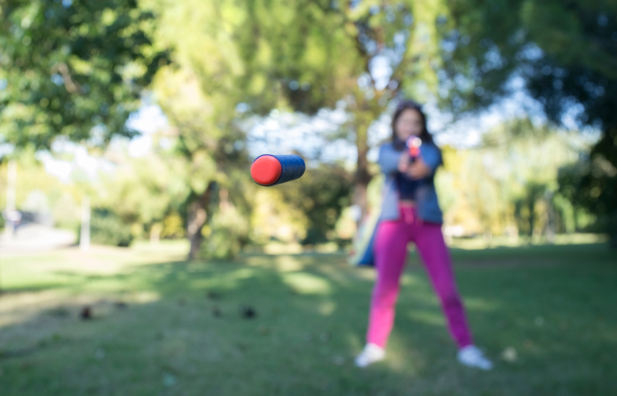 A female aiming a nerf gun with a nerf pellet flying towards the camera