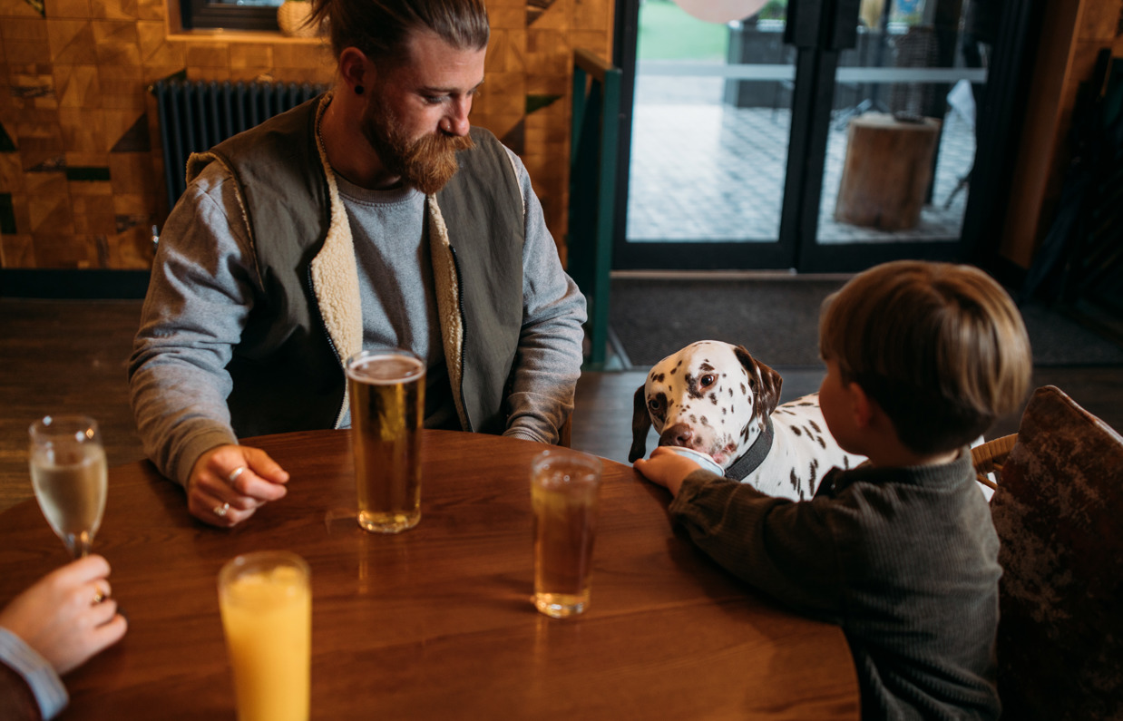 A young boy and his Dad sat at a table in Stargazy Bar & Grill with drinks and feeding a dalmatian dog ice cream