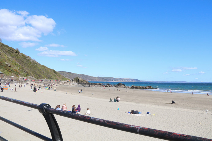A sandy beach with lots of people, blue seas, a promenade and hills in the background
