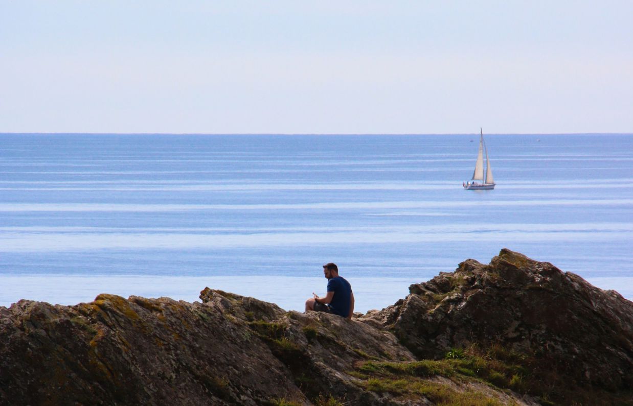 A man sat on the rocky cliffside of the coast with blue sea and a boat in the distance