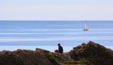 A man sat on the rocky cliffside of the coast with blue sea and a boat in the distance