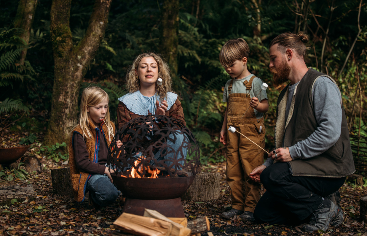 A couple with their two young sons around a fire pit in the woods toasting marshmallows