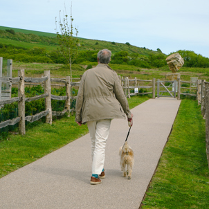 A man walking a dog along a pathway towards a nature trail
